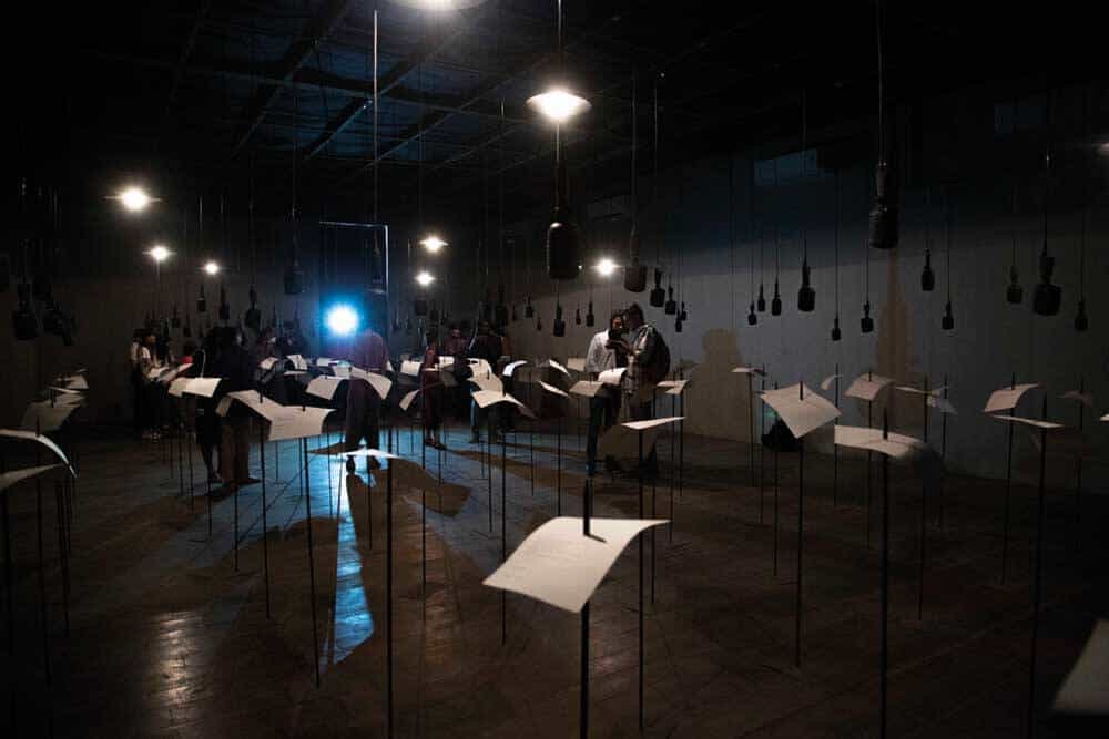 Shilpa Gupta, ‘For, In Your Tongue, I Can Not Fit — 100 Jailed Poets’, Installation view at Kochi-Muziris Biennale 2018, Courtesy of Kochi Biennale Foundation. In this work Shilpa Gupta expands on the artist’s investigations of political borderlines, and how they exist beyond maps to the invisible mechanisms of control and surveillance. The work is an installation of one-hundred speaking microphones that sit above corresponding stakes that each hold a page of poetry. Recitals of a different poet’s work emanate from each microphone in a synchronized chorus. All of the writers who are represented, some living decades or centuries ago, were imprisoned for their poetry or politics, and the installation gives voice to their forced silence. Incarceration instigates a physical boundary between prisoners and the free world. However ‘For, In Your Tongue, I Can Not Fit — 100 Jailed Poets’, points to how orchestrated oppression is harder to detect as it renders those imprisoned voiceless and invisible.