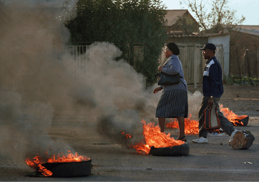 Work, Khumalo Street, 1990. A man and a woman walk through burning barricades to get to work. Thokoza is a small, nondescript township; the main road, Khumalo Street, runs north-south for four kilometres through an elongated triangle from one set of migrant workers’ hostels to another. As the Hostel War cemented frontlines, Khumalo Street became a no-go area, though occasionally we would brave a run along it, sinking low into the car seats while racing through the stop signs and hoping no one would shoot.